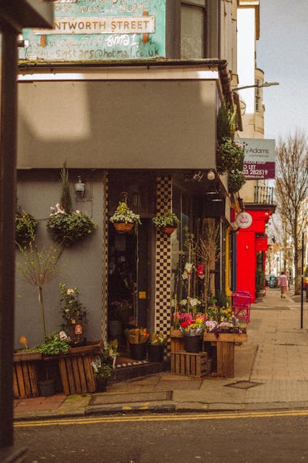 A floral shop on a street corner displays a variety of flower arrangements and potted plants outside its entrance. The display includes colorful bouquets with a mix of roses, daisies, and greenery, wr