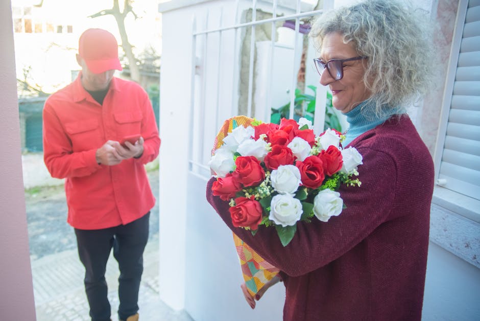 A mature woman with curly gray hair, wearing glasses, a light blue turtleneck, and a maroon cardigan, holds a vibrant bouquet of fresh red and white roses with green foliage, wrapped in colorful patte