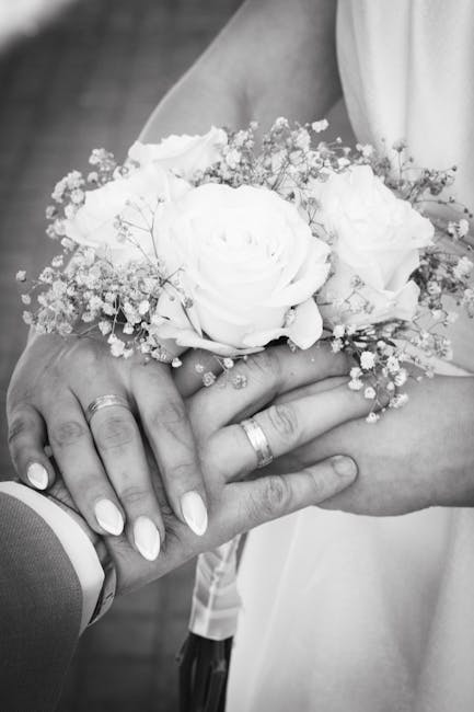 A close-up black-and-white photograph of a bride and groom's hands, the bride wearing a gemstone ring and holding a small bouquet of fresh flowers. The bouquet features large, soft white roses surroun