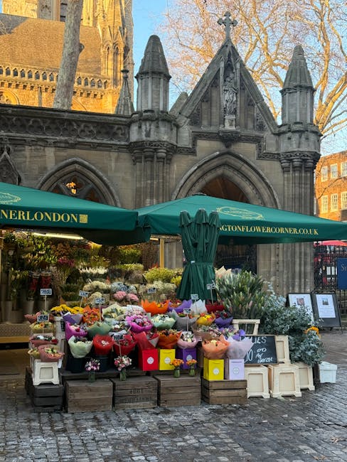 An outdoor flower stall set up in front of an old stone church with Gothic architectural features, including arched windows and pointed spires, during daylight hours. The stall displays a variety of f