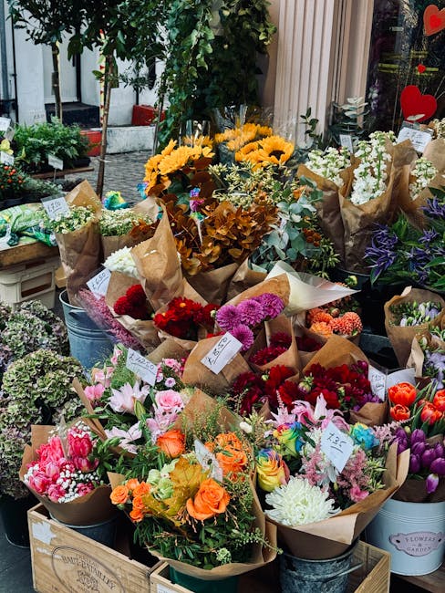 A display of diverse fresh flower bouquets arranged outdoors, each wrapped in brown craft paper and tied with thin ribbons or twine, showcasing a vibrant array of colours including reds, pinks, orange