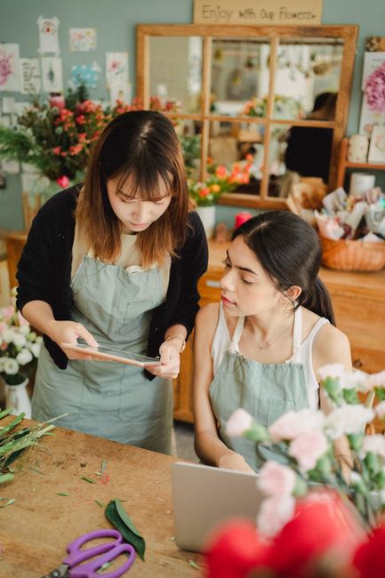 A floral shop scene featuring two women, one standing and the other seated, engaged in arranging and discussing a flower bouquet. The standing woman, with shoulder-length brown hair and wearing a blac