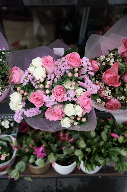 A vibrant floral arrangement featuring a bouquet of fresh pink roses, white carnations, and small pink filler flowers, all artistically wrapped in delicate purple tissue paper with a satin ribbon. The