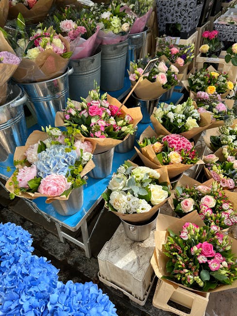 A display of freshly arranged floral bouquets featuring various pastel-colored flowers including pink roses, white lilies, and light blue hydrangeas, wrapped in brown kraft paper and placed in metal b
