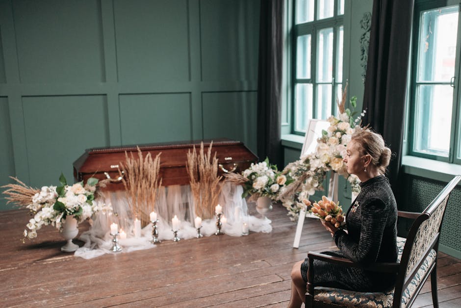 A woman seated on a vintage wooden chair holding a floral arrangement in a black dress, positioned beside a funeral floral display within a room featuring dark green paneled walls and large windows wi
