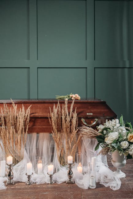 A wooden funeral casket is positioned against a teal-green paneled wall, adorned with a subtle floral arrangement of light pink roses and greenery placed on top. In front of the casket, there are tall